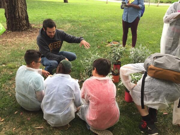 Male graduate student squatting in front of three 5th grade students who are sitting on the ground in front of potted plants