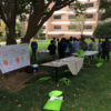 Two tables set under a tree with multiple 5th graders standing around. Against the tree is a banner that reads "Strawberry DNA Extraction Booth. Student faces are covered with smiley faces